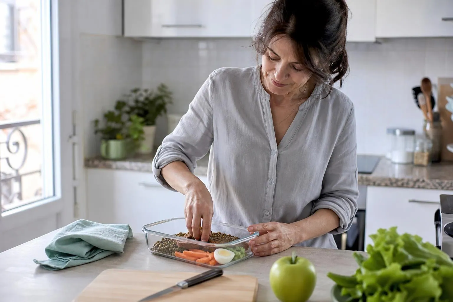 Femme d’une cinquantaine d’années préparant sur un plan de travail clair une boîte repas contenant des lentilles, des crudités et une tranche de pain complet